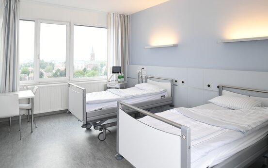 Zweibett-Patientenzimmer mit zwei höhenverstellbaren Betten, Sitzecke, großem Fenster und medizinischer Ausstattung