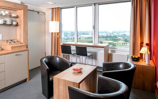 Modern eingerichtete Lounge mit großen Fenstern, drei schwarzen Ledersesseln, Bartisch, Kaffeestation und Ausblick ins Grüne