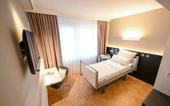 Einzelzimmer in der Klinik mit Krankenbett, Nachttisch, Schreibtisch, Fernseher und viel Tageslicht