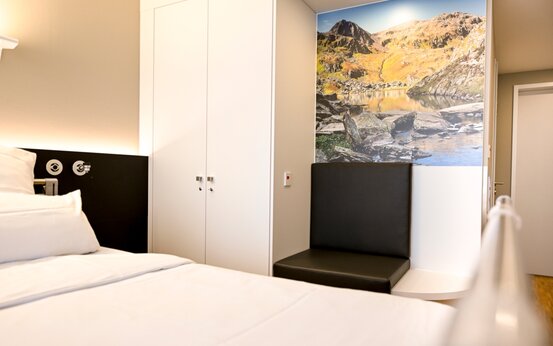 Klinikzimmer mit weißem Bett, großem Kleiderschrank, Sitzgelegenheit und Wandbild einer Berglandschaft