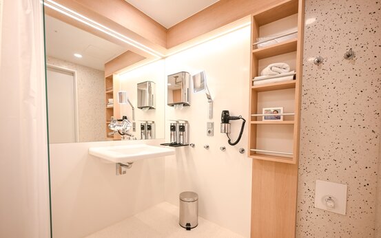 Badezimmer mit unterfahrbarem Waschbecken, großem Spiegel, Seifenspendern, Föhn und Holzregalen mit Handtüchern