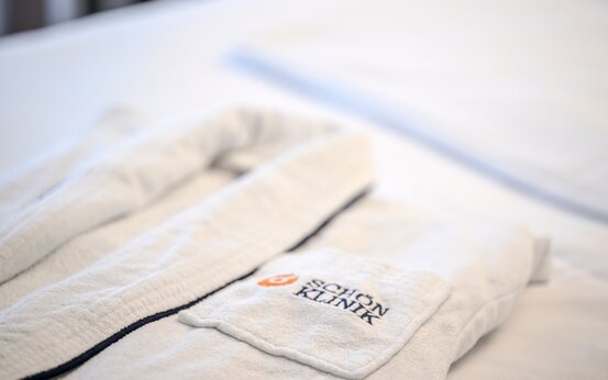 Weißer Bademantel mit Schön Klinik Logo liegt ordentlich auf einem weißen Bett im Wahlleistungszimmer