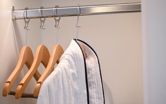 Offener Kleiderschrank mit drei leeren Holzkleiderbügeln und einem weißen Bademantel mit dunkelblauem Rand auf Metallstange
