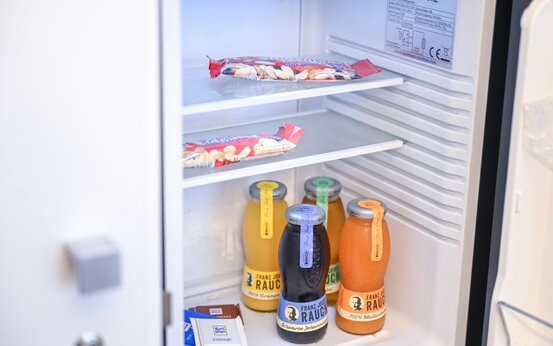 Offener, niedriger Kühlschrank im Klinikzimmer mit Säften, Wasser, Müsliriegeln und Schokolade