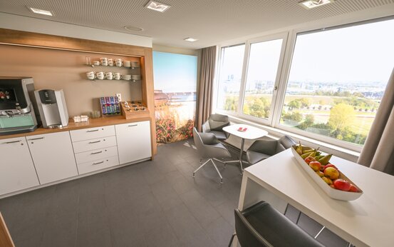 Moderne, helle Klinik-Lounge mit Fensterfront, Sitzgruppen, Stehtisch mit Obstschale und Kaffee-Getränkestation