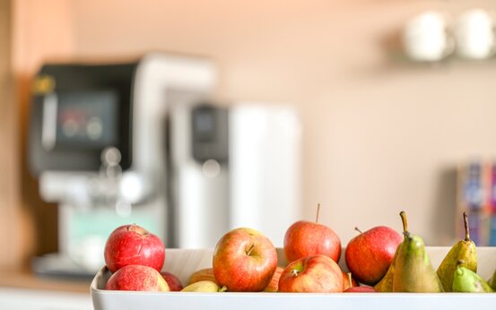 Schale mit frischen Äpfeln und Birnen auf Theke in moderner Klinik-Lounge, dahinter Kaffeevollautomat und Tassenregal