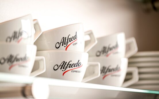 Weiße Espressotassen mit „Alfredo Espresso“-Schriftzug und rotem Akzent, ordentlich gestapelt auf Regal