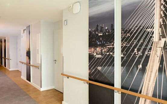 Klinikflur mit Handläufen, hellem Holzfußboden und großformatigen Fototapeten mit Stadt- und Brückenmotiv