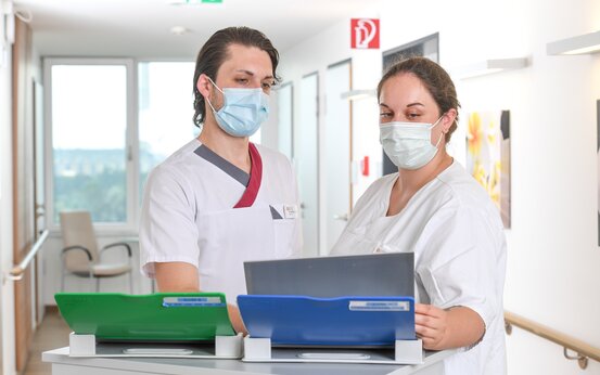 Zwei Pflegekräfte in weißer Arbeitskleidung stehen an einem Stationswagen im hellen Krankenhausflur und betrachten gemeinsam grüne und blaue Aktenordner