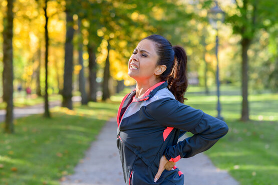 Frau in Sportkleidung hält sich mit schmerzverzerrtem Gesicht den unteren Rücken beim Joggen im herbstlichen Park