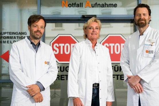 Drei medizinische Fachkräfte in weißen Kitteln vor dem Eingang der Notfallaufnahme mit STOP-Schildern