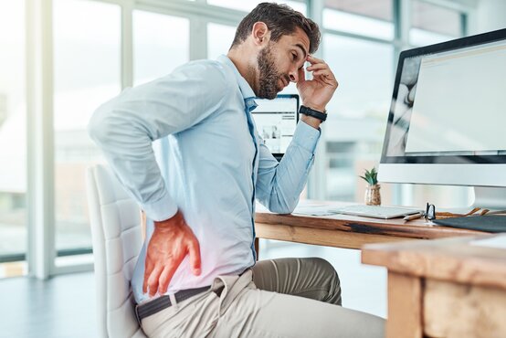 Mann im Business-Outfit hält sich mit schmerzverzerrtem Gesicht den unteren Rücken am Schreibtisch