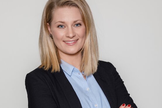 Porträt einer jungen, lächelnden Frau mit blonden, schulterlangen Haaren, schwarzem Blazer und hellblauer Bluse vor grauem Hintergrund