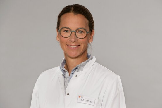 Dr. Tina Maghsoudi. Lächelnde Ärztin mit Brille im weißen Kittel und Namensschild vor neutralem Hintergrund