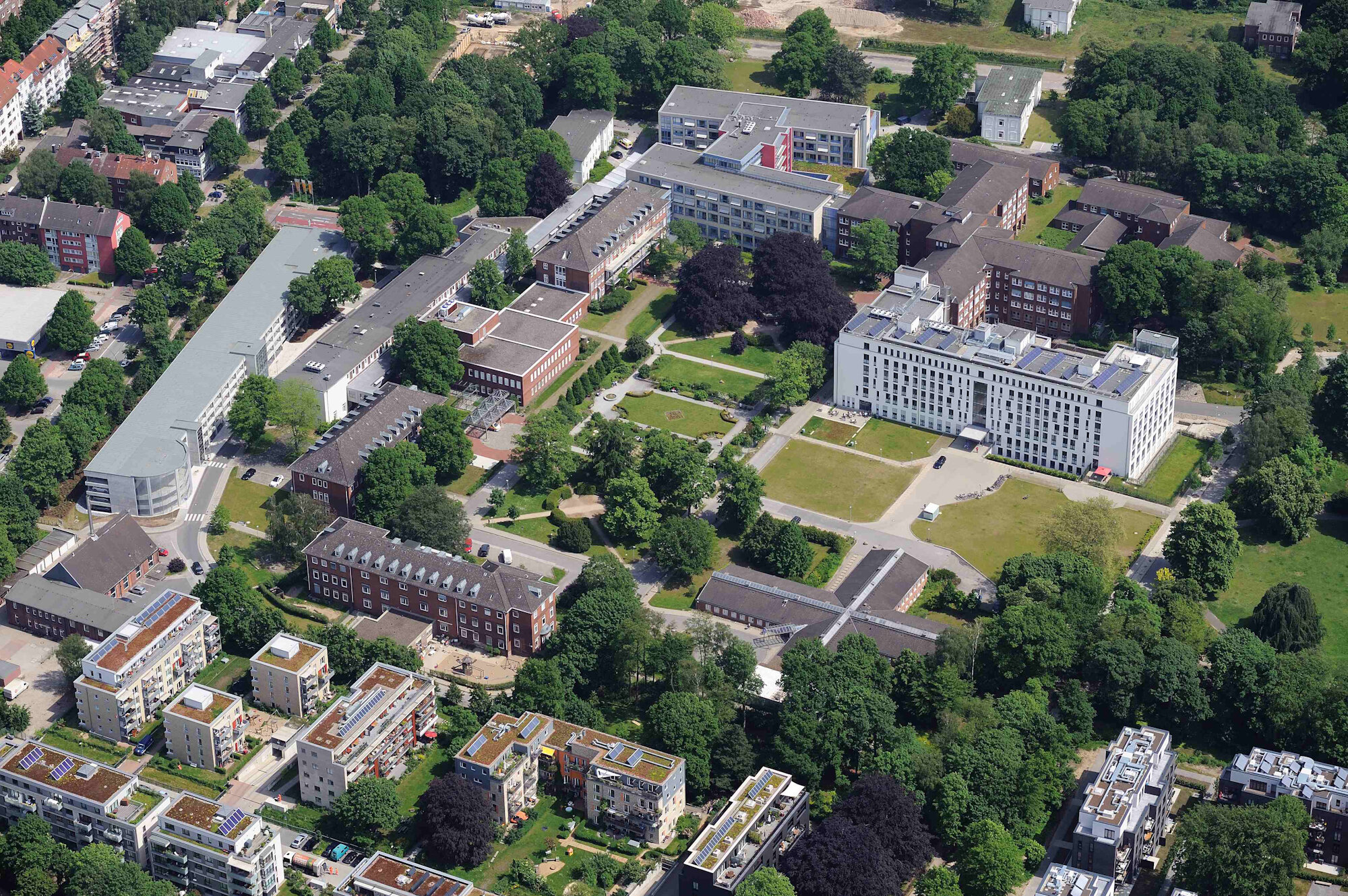 Luftaufnahme eines modernen Klinikgeländes mit weißen Gebäuden, Grünflächen, Bäumen und großem Vorplatz