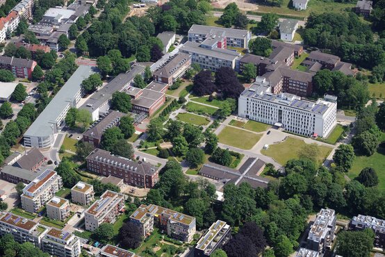 Luftaufnahme eines modernen Klinikgeländes mit weißen Gebäuden, Grünflächen, Bäumen und großem Vorplatz