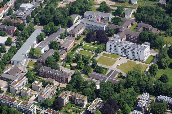 Luftaufnahme eines modernen Klinikgeländes mit weißen Gebäuden, Grünflächen, Bäumen und großem Vorplatz