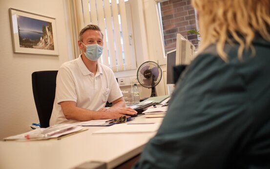 Männlicher Arzt mit Mund-Nasen-Schutz berät Patientin in heller Adipositas-Sprechstunde, Unterlagen auf dem Tisch