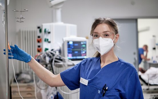 Krankenhaus-Mitarbeiterin in blauer OP-Kleidung, Schutzbrille und FFP2-Maske am Infusionsständer