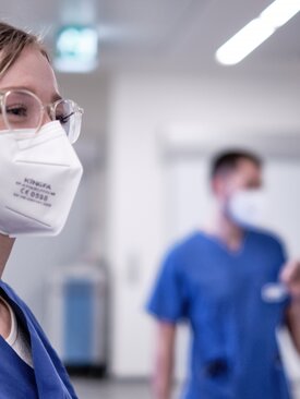 Junge Pflegekraft mit FFP2-Maske und Brille im Krankenhausflur, zwei Kollegen im Hintergrund