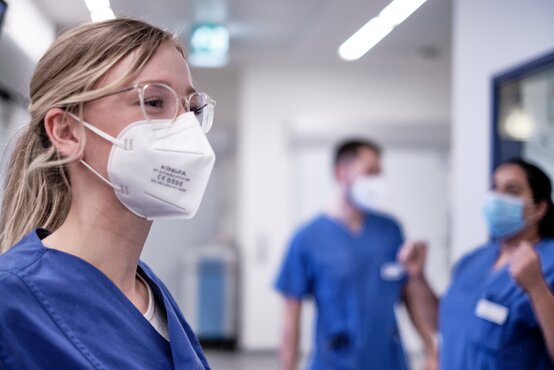 Junge Pflegekraft mit FFP2-Maske und Brille im Krankenhausflur, zwei Kollegen im Hintergrund