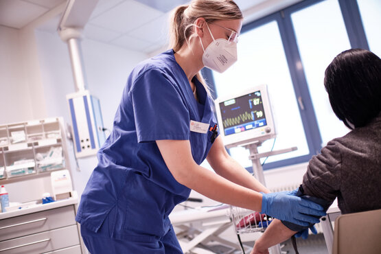 Medizinische Fachkraft in blauer Kleidung, FFP2-Maske und Schutzbrille misst Blutdruck bei Patientin