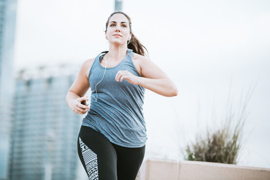 Junge Frau joggt im Freien in Sportkleidung mit Kopfhörern und Smartphone, unscharfe Gebäude im Hintergrund