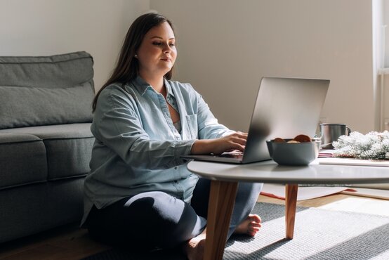 Junge Frau sitzt im modernen Wohnzimmer auf dem Boden, arbeitet am Laptop, Kekse und Tasse auf Couchtisch, Sofa im Hintergrund