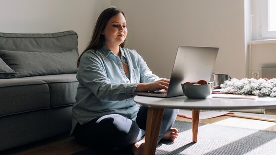 Junge Frau sitzt im modernen Wohnzimmer auf dem Boden, arbeitet am Laptop, Kekse und Tasse auf Couchtisch, Sofa im Hintergrund