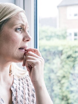 Frau mit blonden Haaren steht besorgt am Fenster und schaut raus