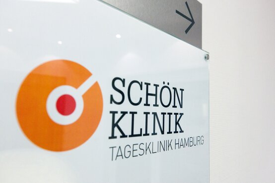 Eingangsschild der Schön Klinik Tagesklinik Hamburg aus Glas mit orange-rotem Logo