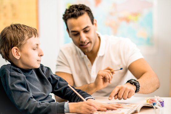 Therapeut unterstützt Jungen im Rollstuhl bei motorischer Übung am Tisch