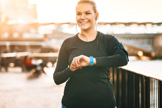 Junge Frau joggt lachend in Sportkleidung am Fluss, prüft Fitnessuhr, Bewegung und Lebensfreude