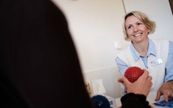 Physiotherapeutin übergibt Patientin in der Klinik einen roten Igelball zur Handrehabilitation, blauer Ball liegt auf dem Tisch