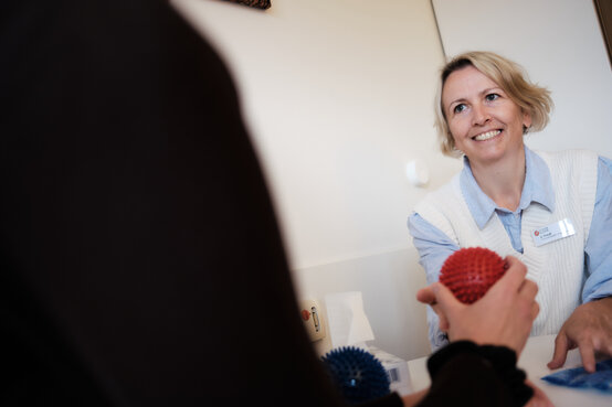 Physiotherapeutin übergibt Patientin in der Klinik einen roten Igelball zur Handrehabilitation, blauer Ball liegt auf dem Tisch