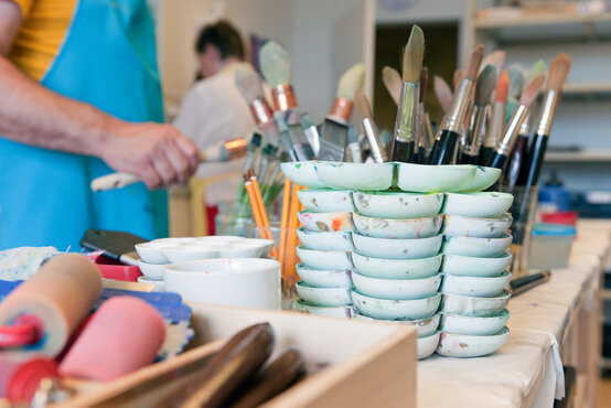 Kunsttherapie-Raum in Klinik mit vielen Pinseln, Farbpaletten und Malutensilien, Patient und Therapeutin