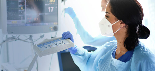 Junge Frau in Schutzkleidung und FFP2-Maske bedient Überwachungsmonitor auf Intensivstation, Vitalparameter sichtbar