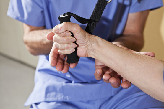 Arzt  in blauer Kleidung unterstützt ältere Person bei Hand- und Armrehabilitation mit Trainingsgriff