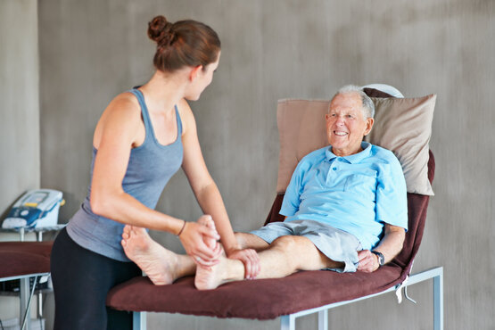 Physiotherapeutin hilft älterem Mann bei Dehnübungen auf Behandlungsliege, beide lächeln