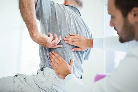 Mediziner untersucht Rücken eines älteren Mannes mit Rückenschmerzen, Patient zeigt auf schmerzende Lendenwirbelsäule