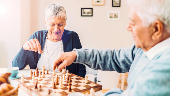 Ältere Frau und Mann spielen lächelnd Schach am Tisch, Kaffee und Croissants 