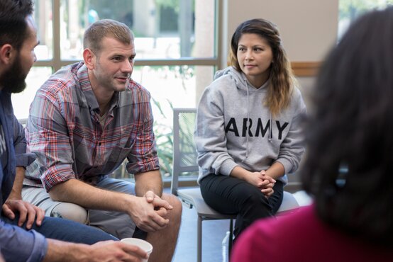 Erwachsene sitzen in Stuhlkreis in hellem Raum, Mann im karierten Hemd und Frau im „ARMY“-Hoodie hören aufmerksam zu, Gruppentherapie