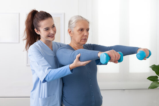 Lächelnde junge Krankenschwester, die einer älteren Frau während einer Physiotherapie-Sitzung in einem Pflegeheim mit leichten Hantelübungen hilft. Der ältere Patient sieht glücklich und engagiert aus, Unterstützung in der Gesundheitsversorgung.