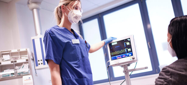 Krankenschwester im blauen Kittel mit Maske erklärt Patientin im Klinikzimmer die Werte auf einem Monitor