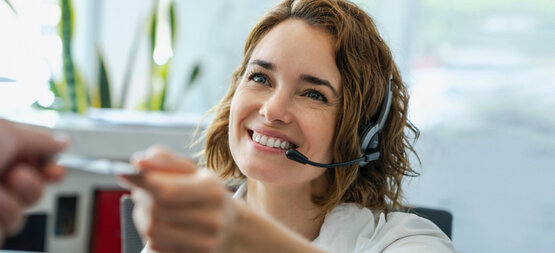 Lächelnde Frau mit Headset am Empfang reicht freundlich eine Karte, helle Büroräume und grüne Pflanzen im Hintergrund