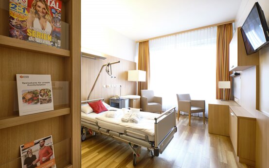 Modernes, helles Krankenhauszimmer mit Einzelbett, rotem Kissen, Holzmöbeln, zwei Sesseln und großem Fenster mit Vorhängen
