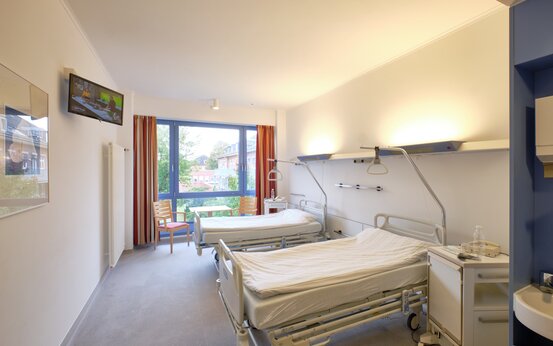 Helles, modernes Krankenzimmer mit zwei weißen Krankenhausbetten, Nachttisch, Fernseher, großem Fenster und Holzstuhl