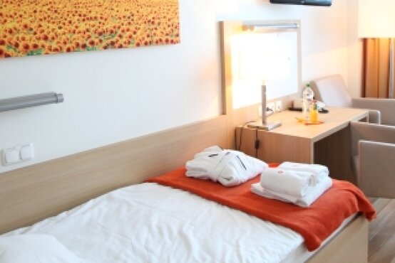 Modernes Einzelbett mit weißer Bettwäsche und oranger Decke in hellem, barrierefreiem Hotelzimmer mit Sitzbereich