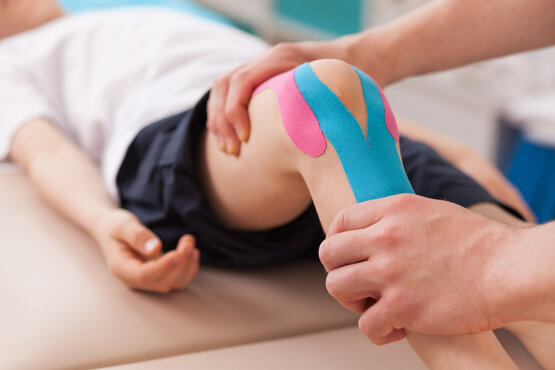 Kind liegt auf Behandlungsliege, Therapeut bringt blau-pinkes Kinesio-Tape am Knie an