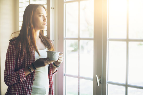 Junge Frau mit langen braunen Haaren steht nachdenklich mit Kaffeetasse am Fenster im Sonnenlicht